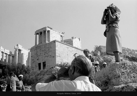 Acropole, Athènes, Grèce, 1979, photographie de Jean pierre Morcrette. La photo, en noir et blanc, montre, vestiges, colonnes, constructions en pierre et touristes. Un homme photographie le site, tandis qu’une femme sur un rocher prend une photo dans la direction opposée. D’autres visiteurs sont dispersés dans la scène.