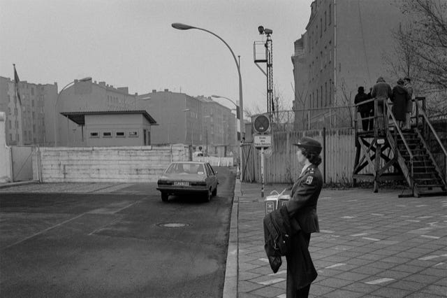 Berlin-Ouest, poste-frontière Chausseestraße vers Berlin-Est, RFA, 1984*, photographie de Jean pierre Morcrette. Une rue avec une voiture qui s’apprête à passer la frontière ; une femme militaire tenant un paquet debout sur le trottoir ; un mur en arrière-plan ; cinq personnes sur une plateforme regardent de l’autre côté du poste-frontière. Un panneau en allemand « Ende des franz Sektors » : Fin du secteur français.