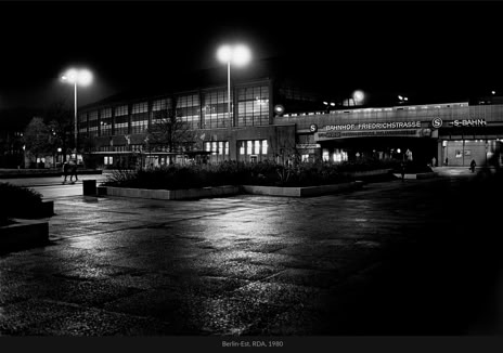 Berlin-Est, RDA, 1980, photographie de Jean pierre Morcrette. Paysage urbain nocturne en noir et blanc devant la gare Friedrichstrasse à Berlin-Est. Des lampadaires éclairent une allée mouillée. Quelques personnes près de l’entrée, avec des plantes en bacs.