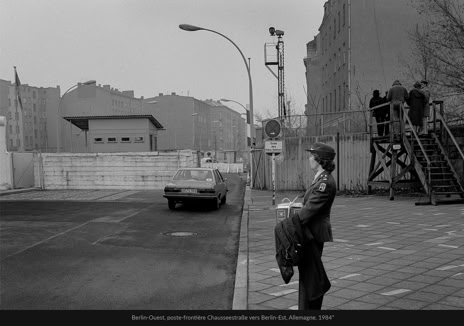 Berlin-Ouest, poste-frontière Chausseestraße vers Berlin-Est, RFA, 1984*, photographie de Jean pierre Morcrette. Une rue avec une voiture qui s’apprête à passer la frontière ; une femme militaire tenant un paquet debout sur le trottoir ; un mur en arrière-plan ; cinq personnes sur une plateforme regardent de l’autre côté du poste-frontière. Un panneau en allemand « Ende des franz Sektors » : Fin du secteur français.